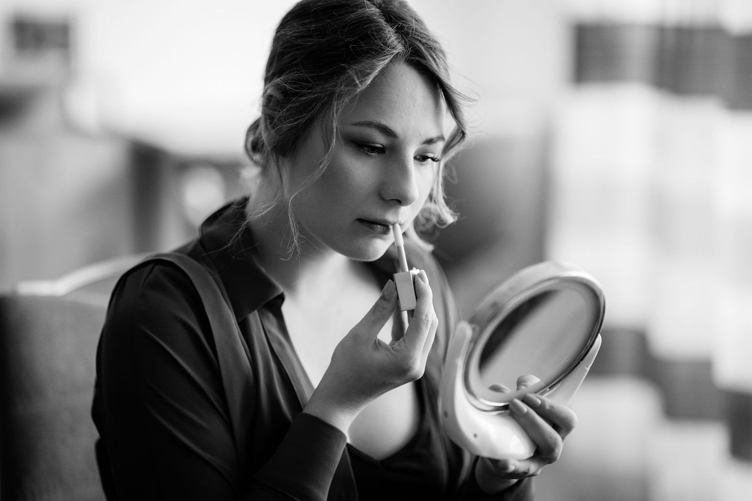 Black and white photo of a young woman applying makeup with a brush while looking into a small round mirror.