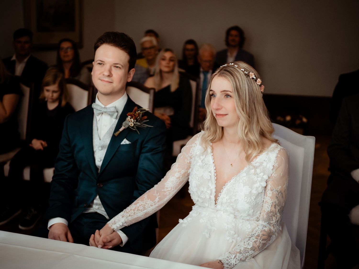 Bride and groom holding hands at wedding ceremony altar, bride in lace gown, groom in teal suit.