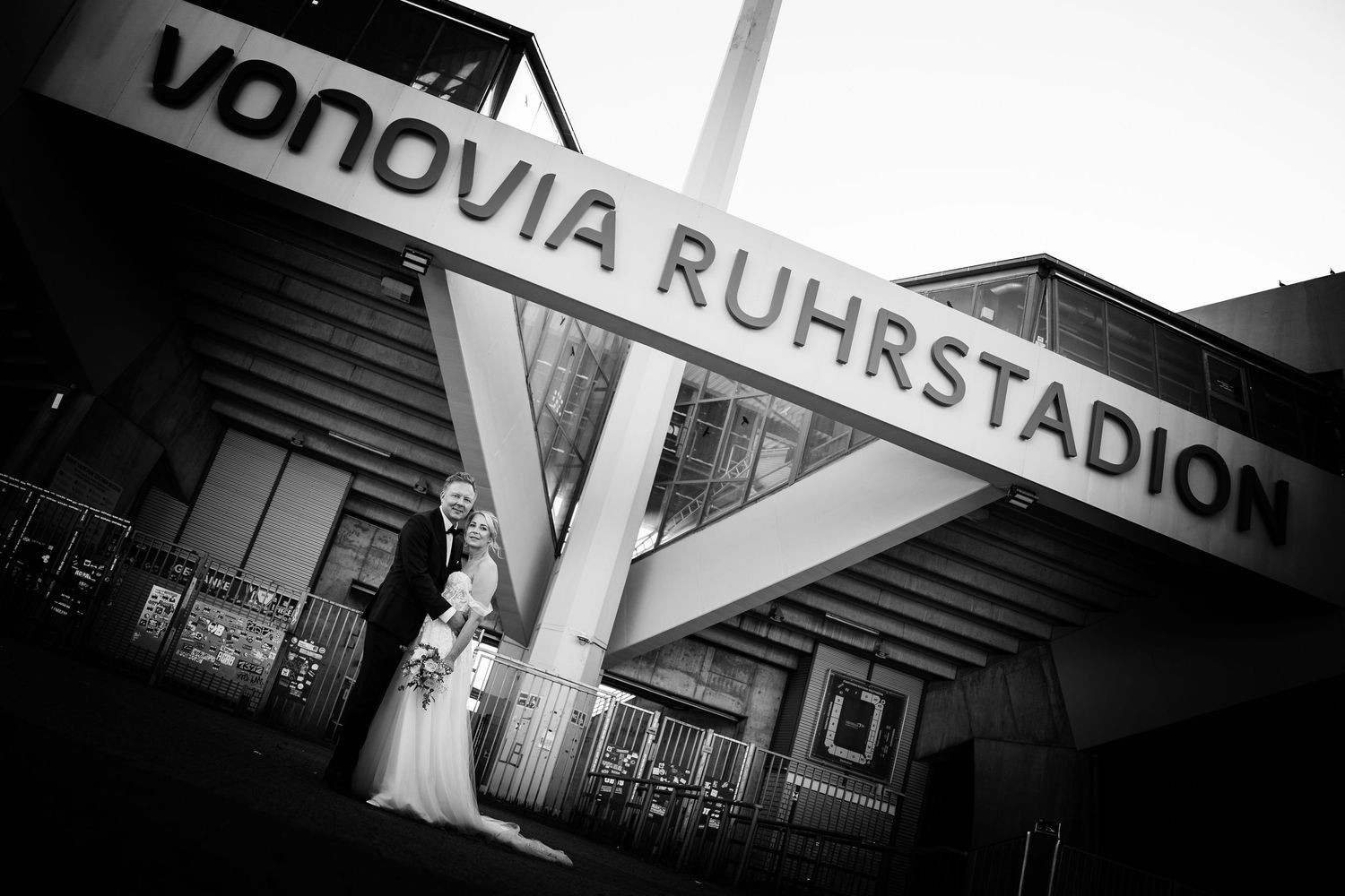 Black and white photo of bride and groom posing at Vonovia Ruhrstadion stadium entrance.