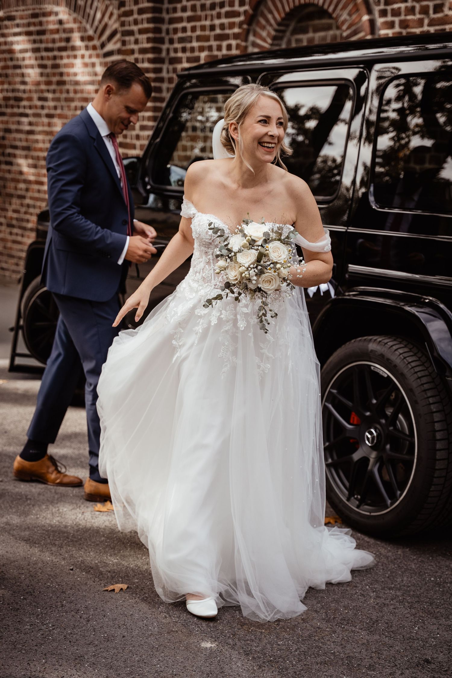 Smiling bride in off-shoulder lace gown holding white bouquet stepping out of black SUV.