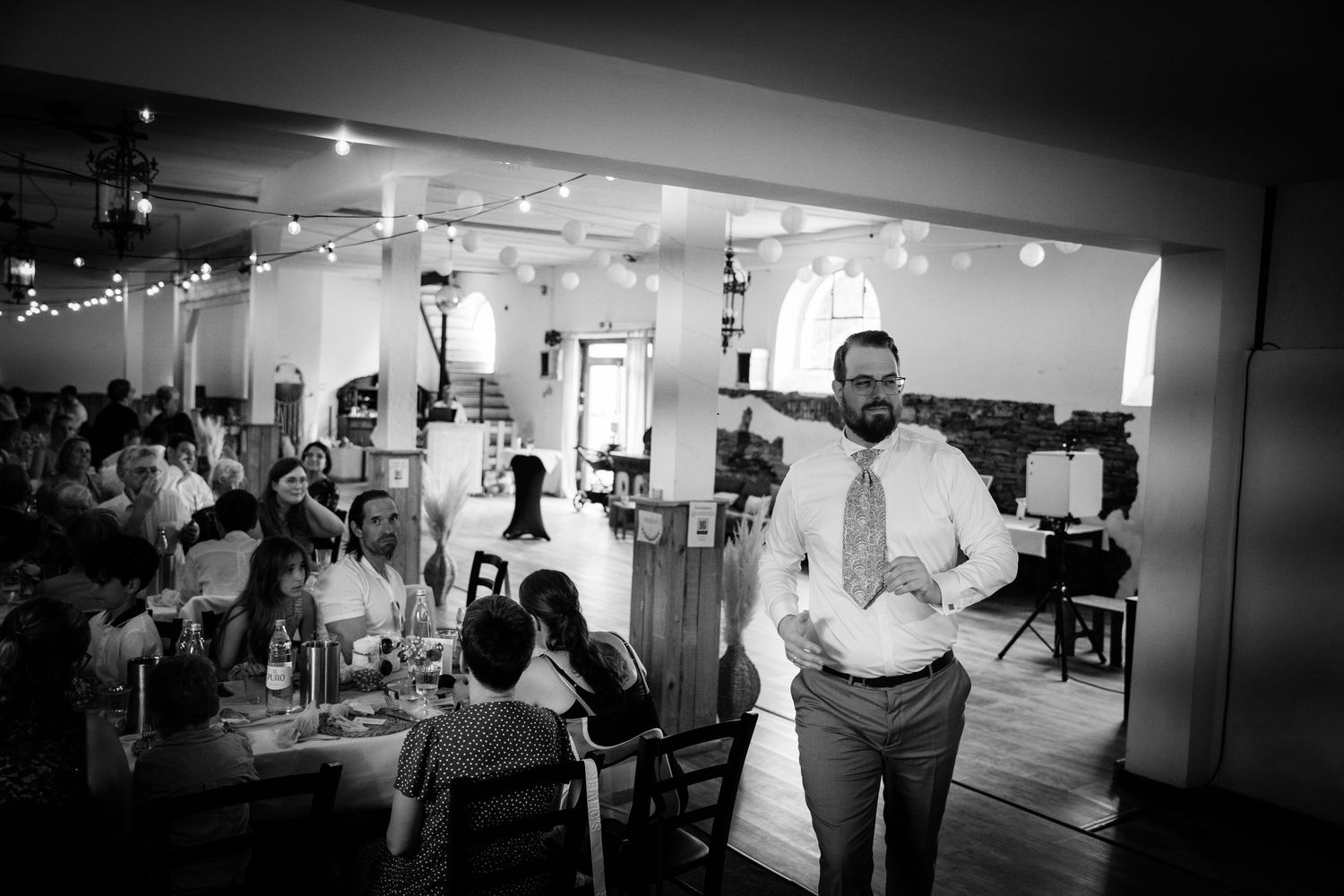 Black and white photo of a man in suit standing before a bright reception hall full of guests.
