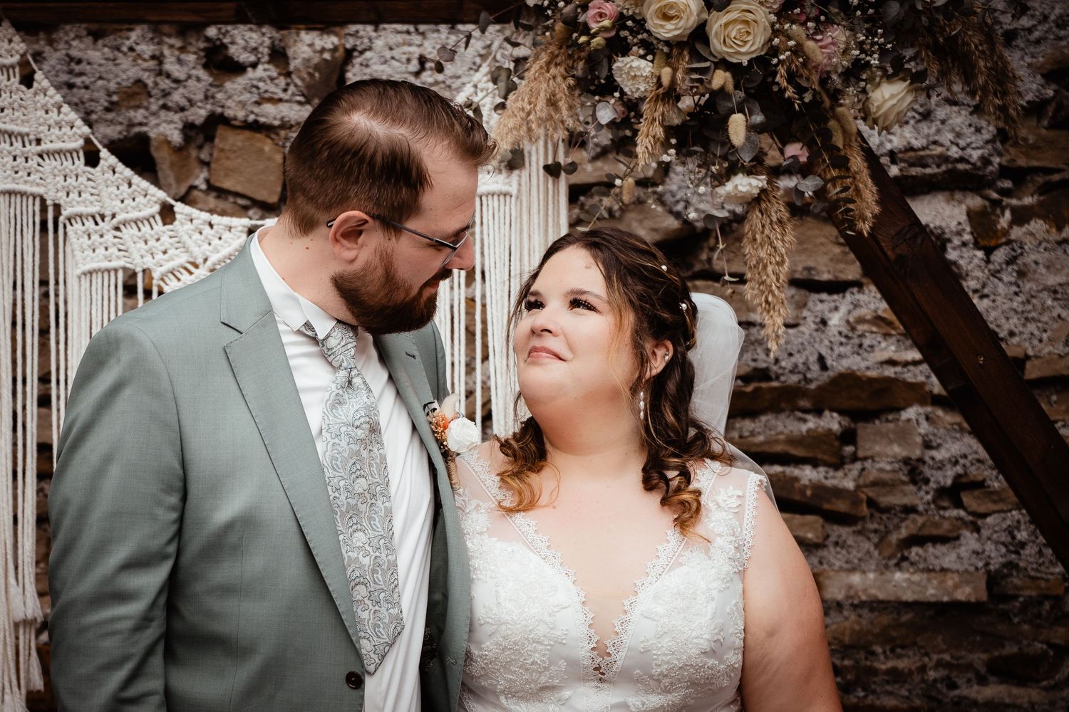 Bride and groom share an intimate glance before a macrame backdrop adorned with dried floral arrangements.