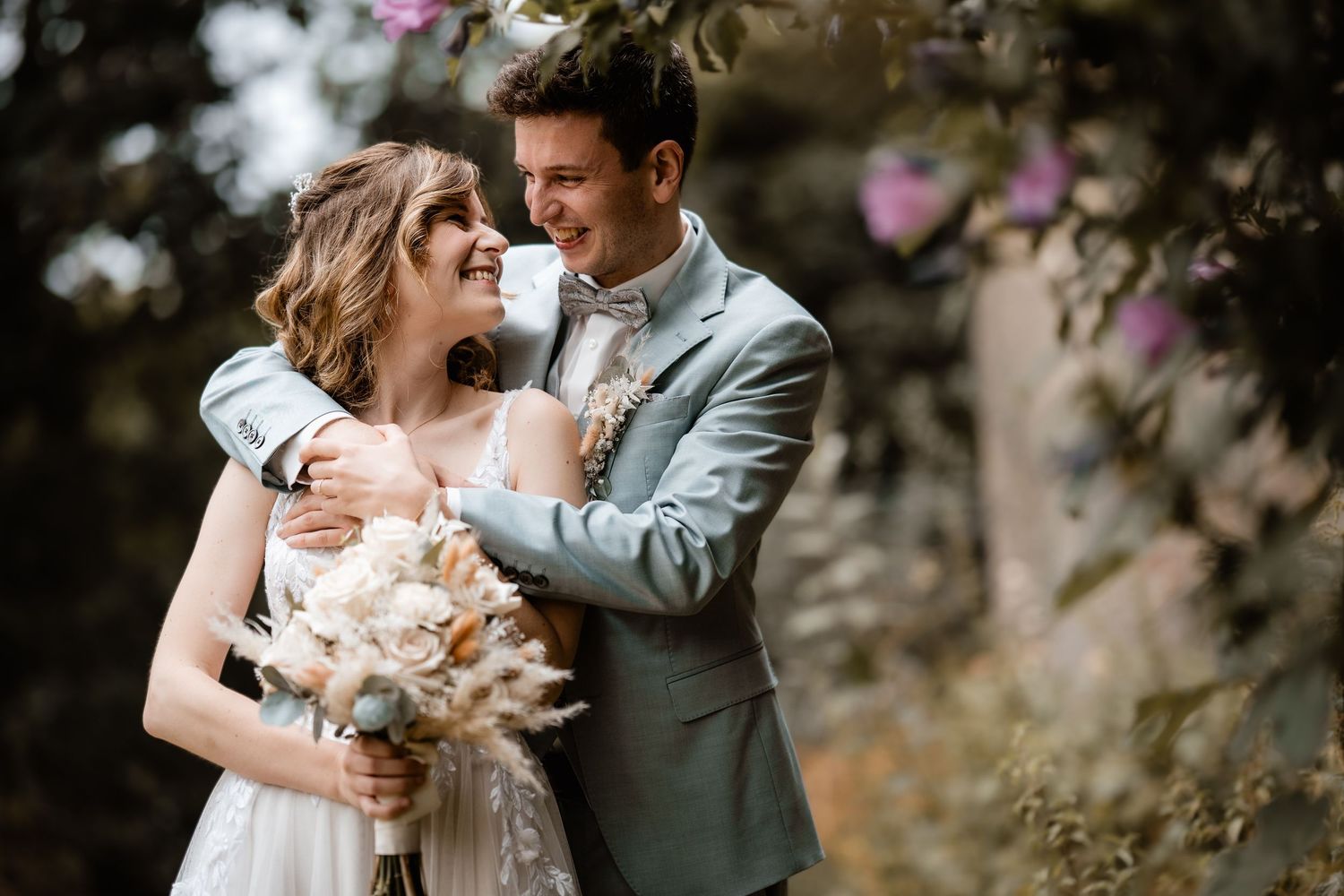 Happy newlyweds embrace outdoors, bride holding white bouquet, groom in gray suit, surrounded by blooming pink flowers.