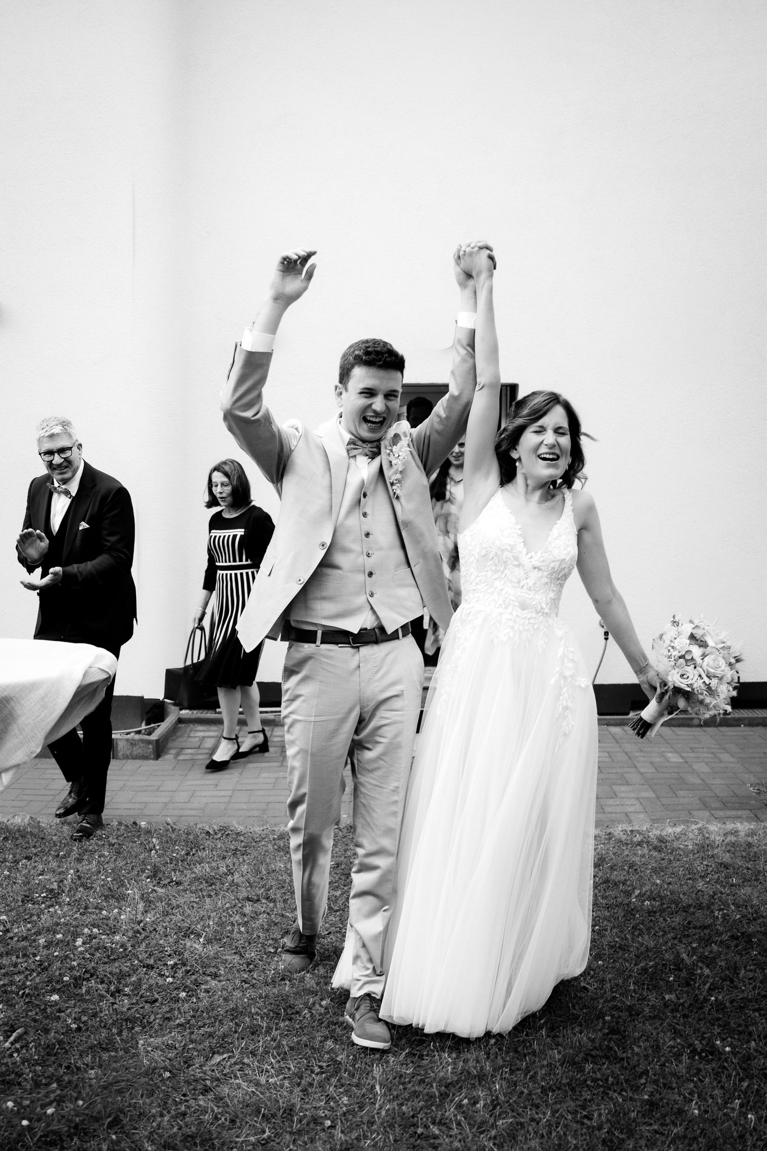 Joyful newlyweds raise hands in celebration after wedding ceremony outdoors.