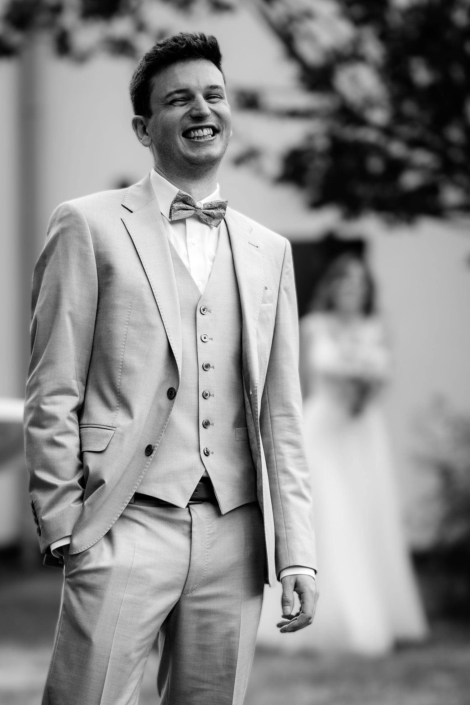 A smiling groom in a light gray three-piece suit with bow tie laughs joyfully on his wedding day.