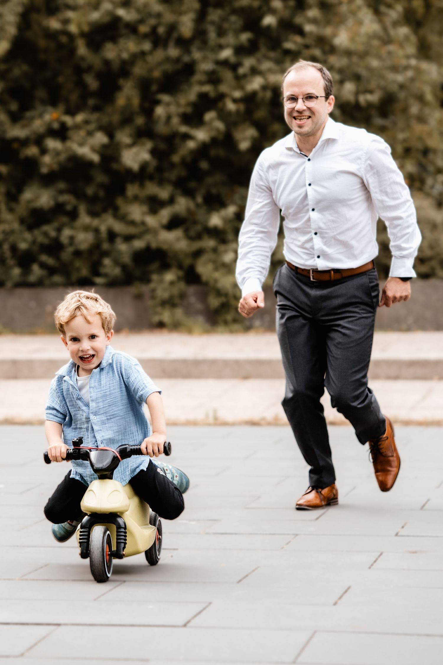 Smiling man chases a laughing young boy riding a toy scooter on a paved outdoor path.