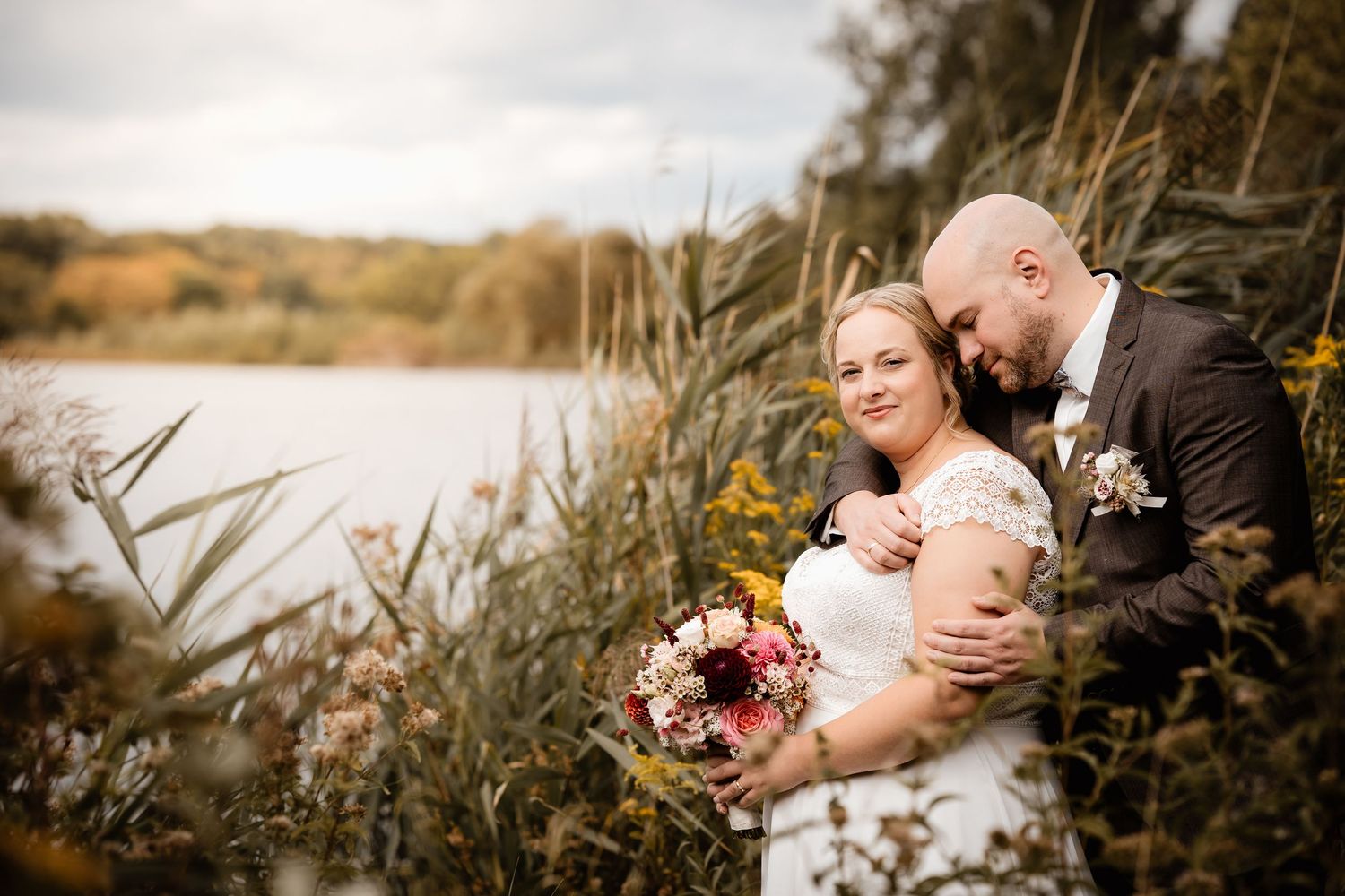 Bride and groom embracing near a lakeside with autumn foliage, bride holding a colorful bouquet.
