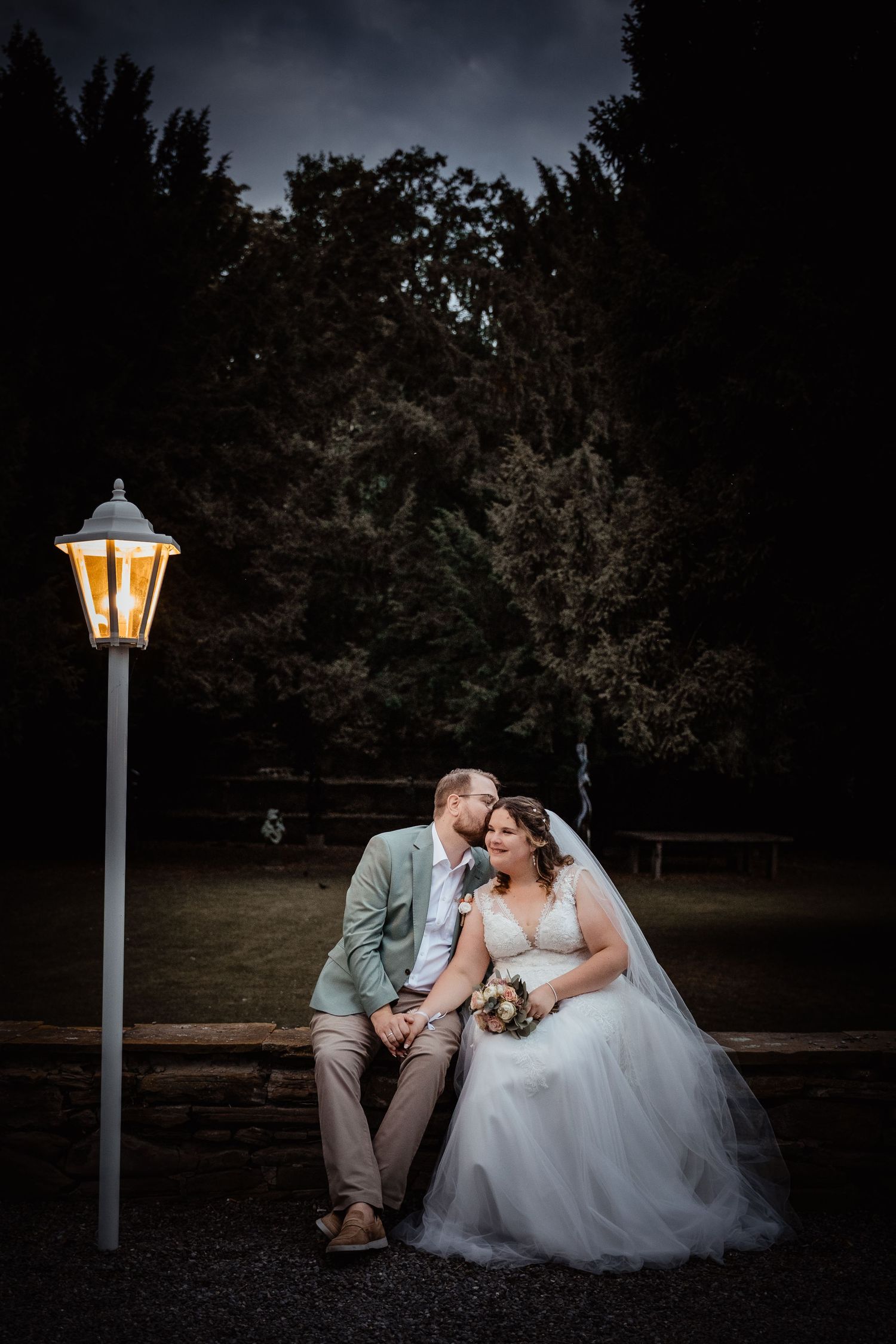 Bride and groom sharing romantic kiss by glowing lantern on bench in moody nighttime garden setting.