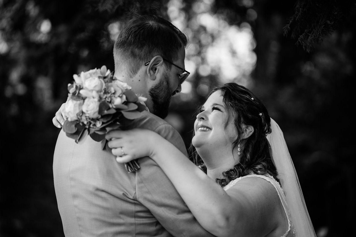 Black and white photo of smiling bride embracing groom who holds a floral bouquet under lush trees.