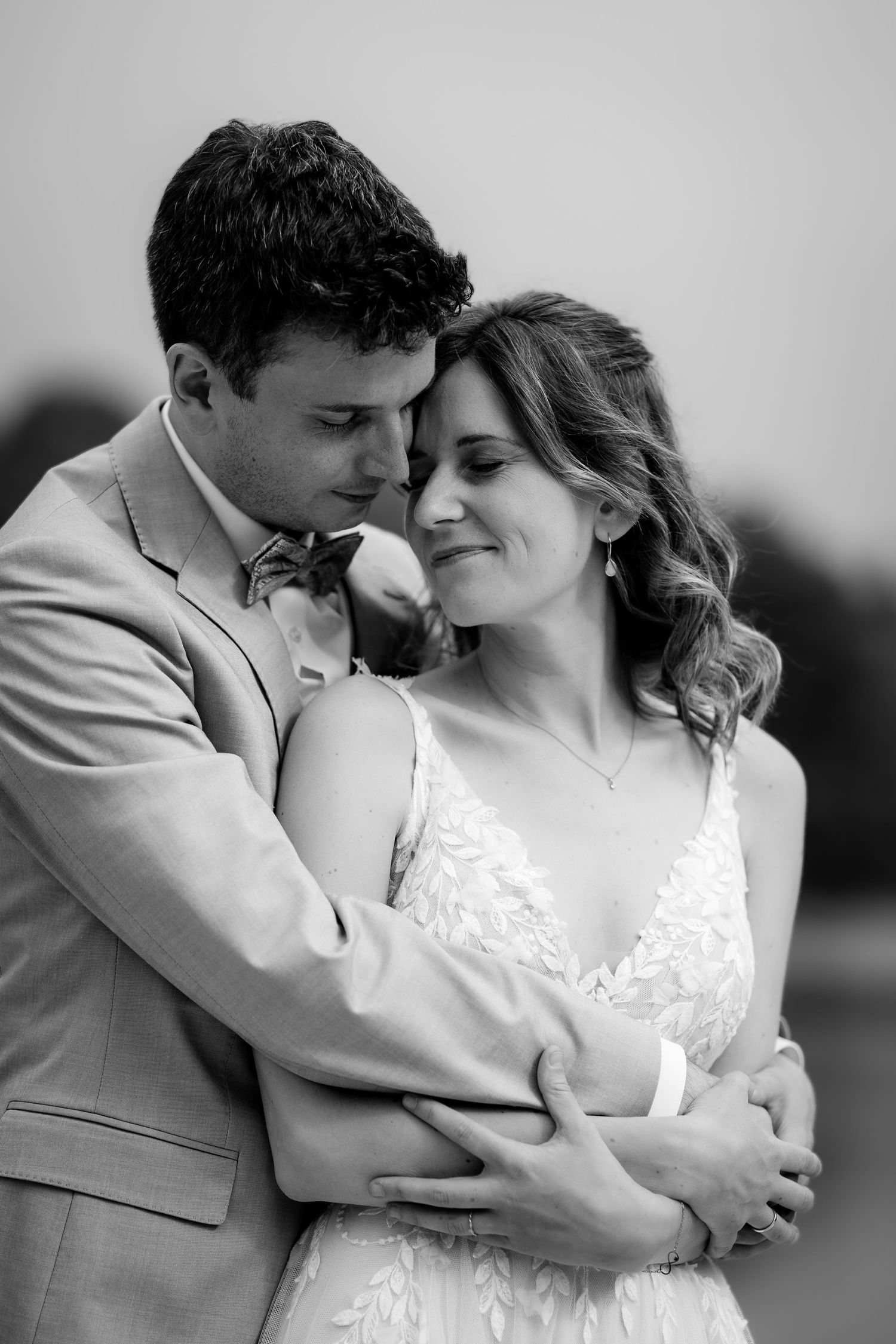 Black and white wedding photo of groom embracing bride from behind, both smiling softly in elegant attire.