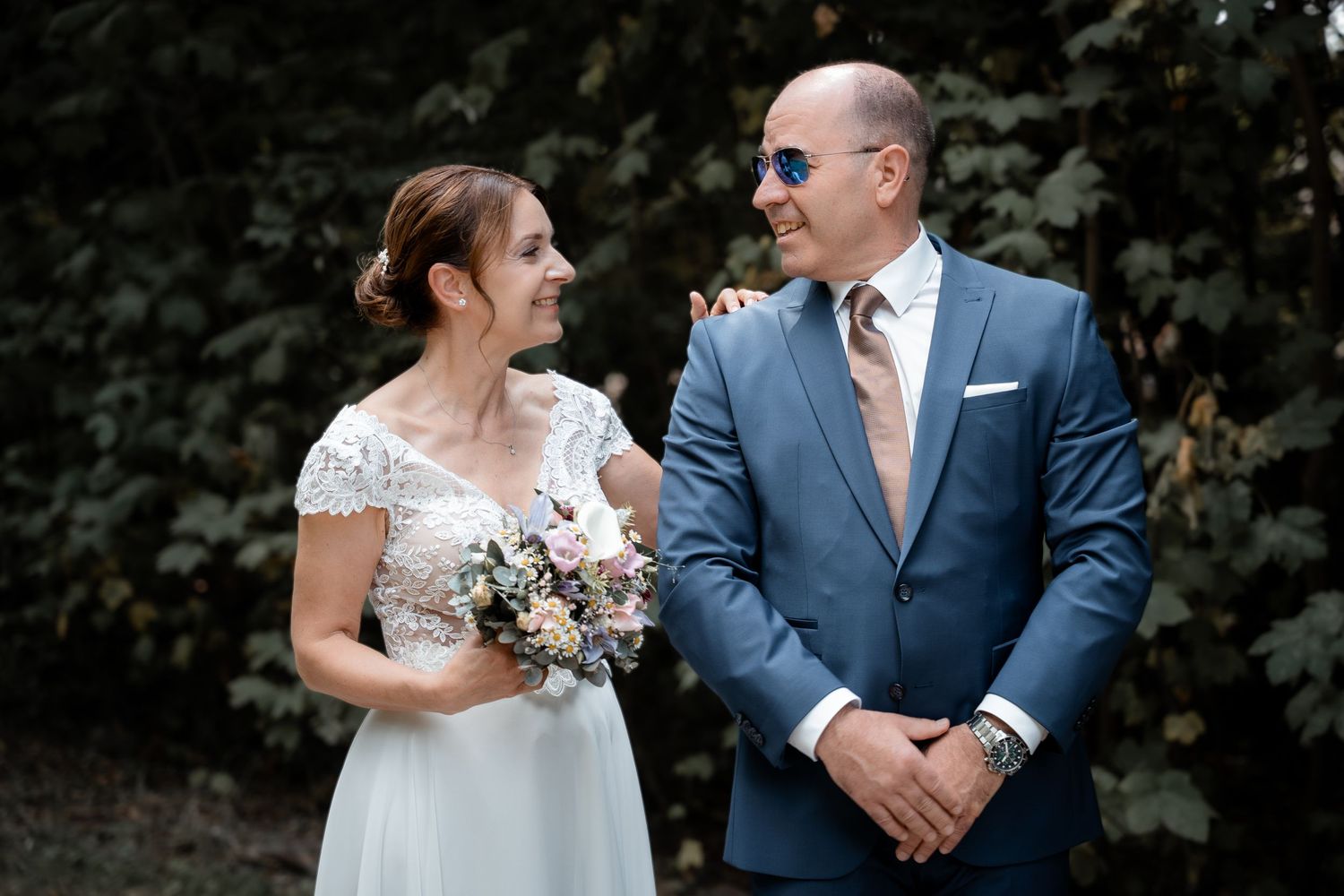 Bride and groom smiling at each other outdoors, she holds a bouquet, he wears a blue suit with sunglasses.