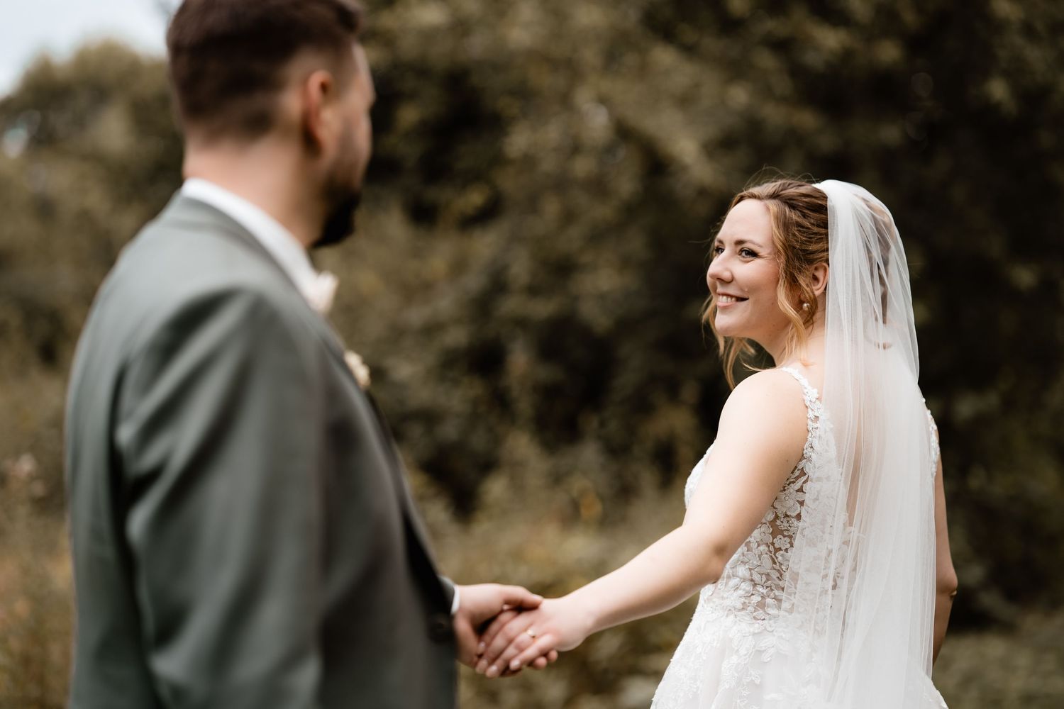 Bride in lace gown and veil smiles at groom in gray suit while holding hands outdoors at wedding.