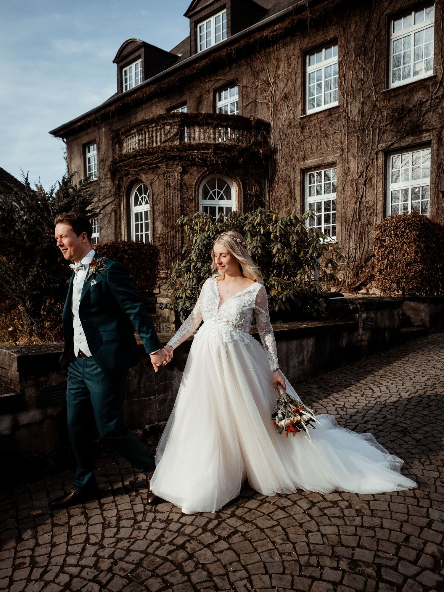 Bride and groom walk hand-in-hand on cobblestones before a grand ivy-covered stone manor on their wedding day.