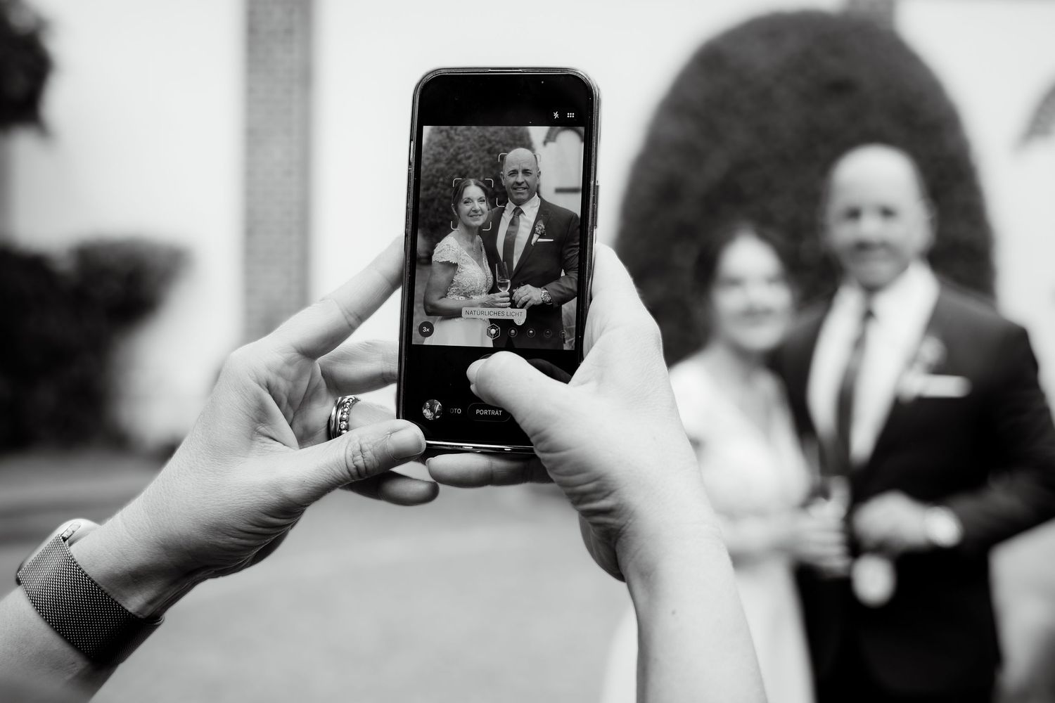 Person photographing a couple in formal attire on a smartphone at a wedding event, black and white photo.