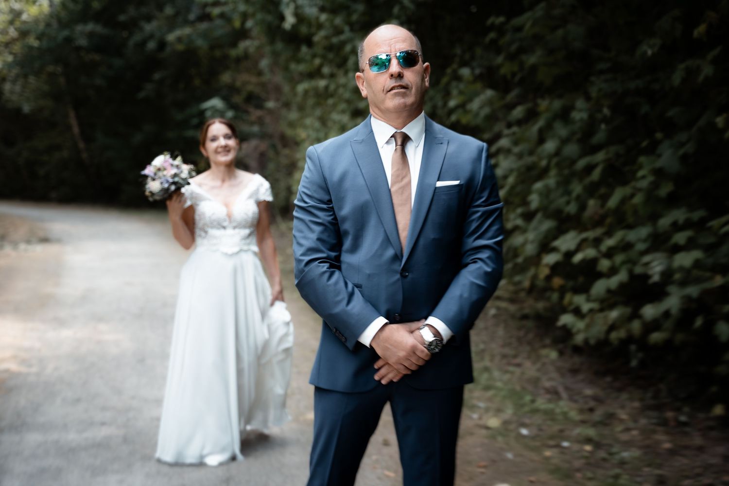 Bride in white gown holding bouquet stands behind groom in blue suit with sunglasses on a tree-lined path.