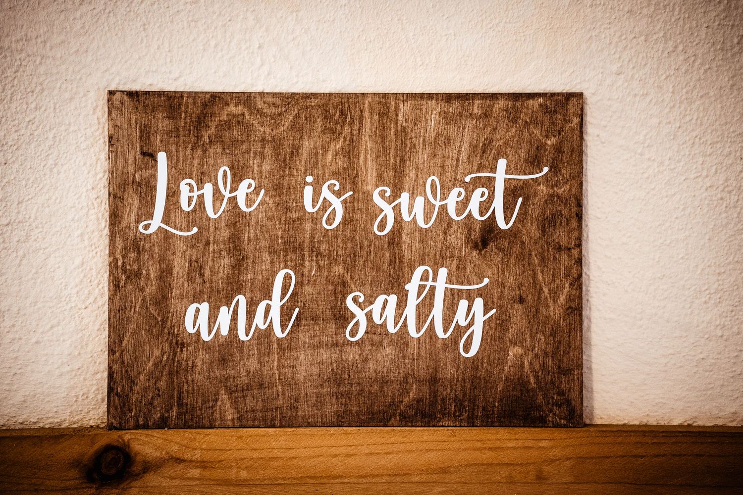 Rustic wooden sign reading Love is sweet and salty leaning against a white wall.