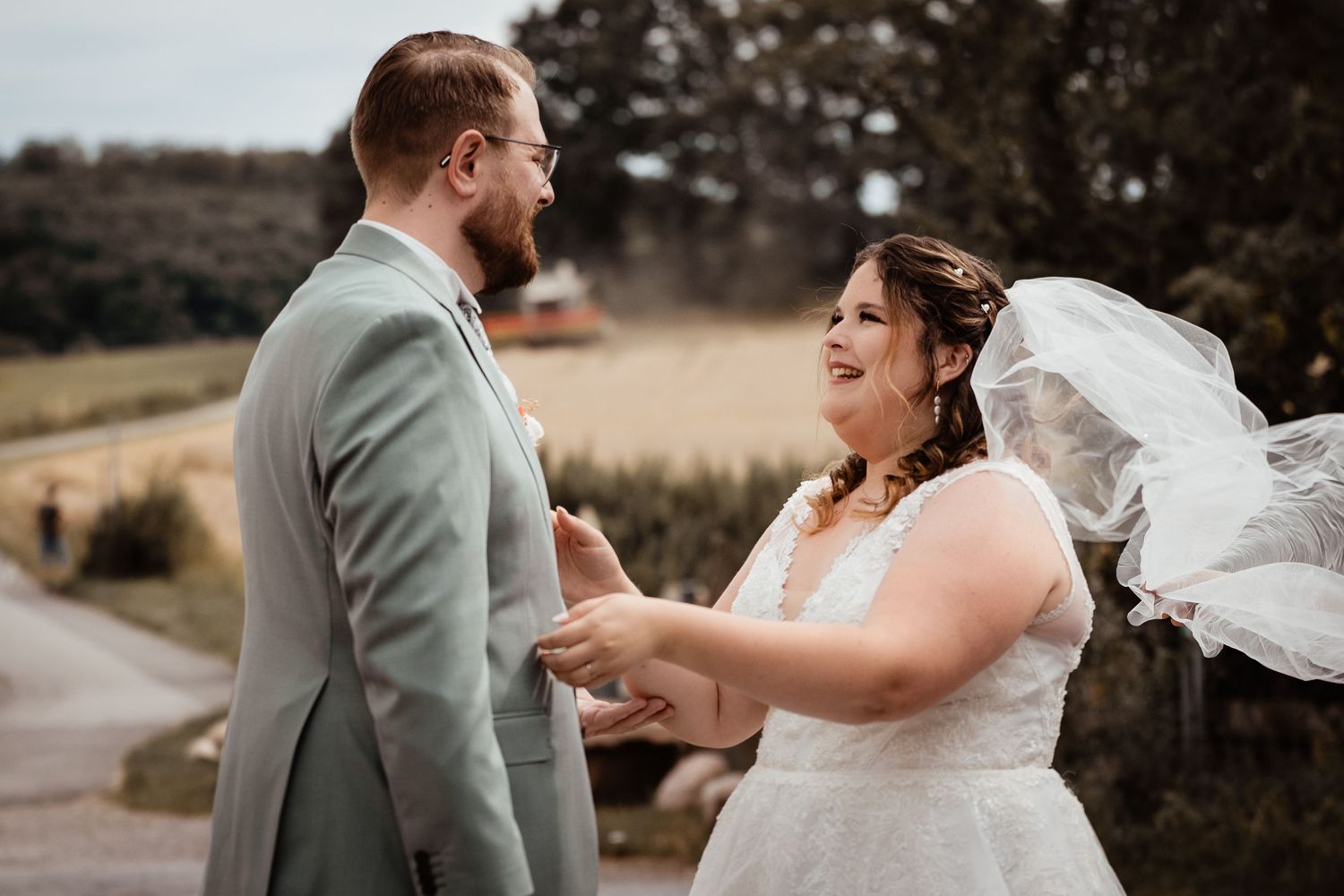 A laughing bride in a white lace gown with flowing veil holds hands with groom in sage green suit outdoors.