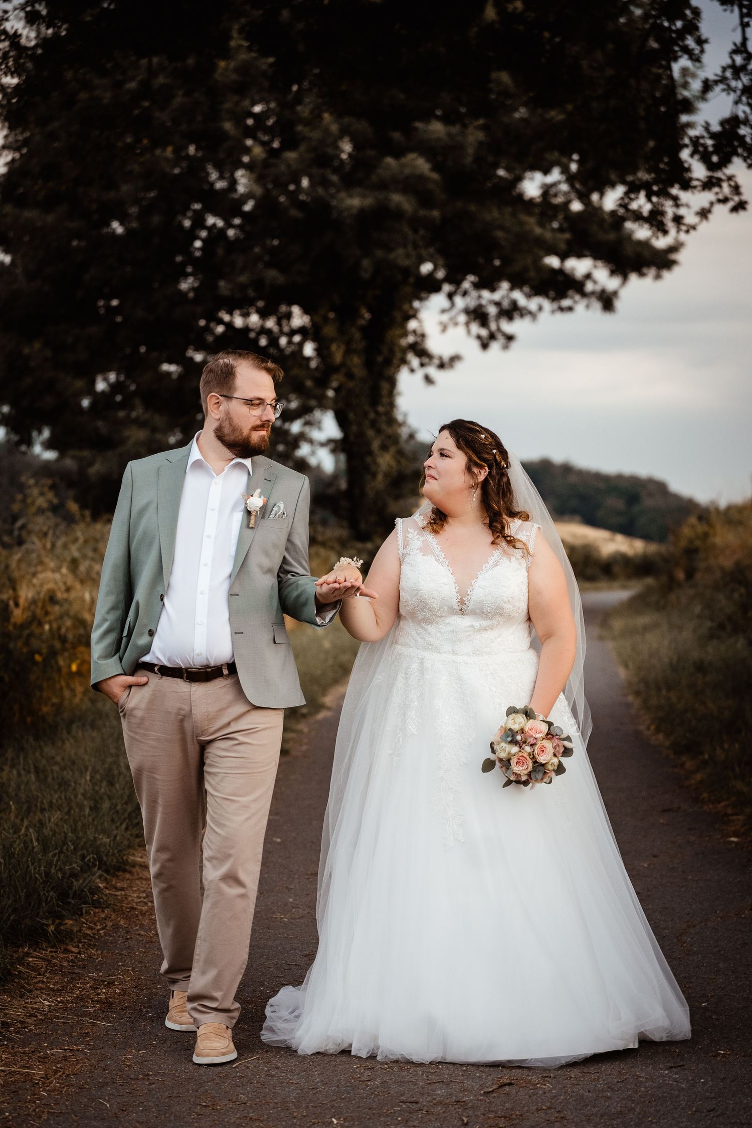 Bride and groom walk hand-in-hand on a scenic country road, she in a white gown, he in a sage blazer.