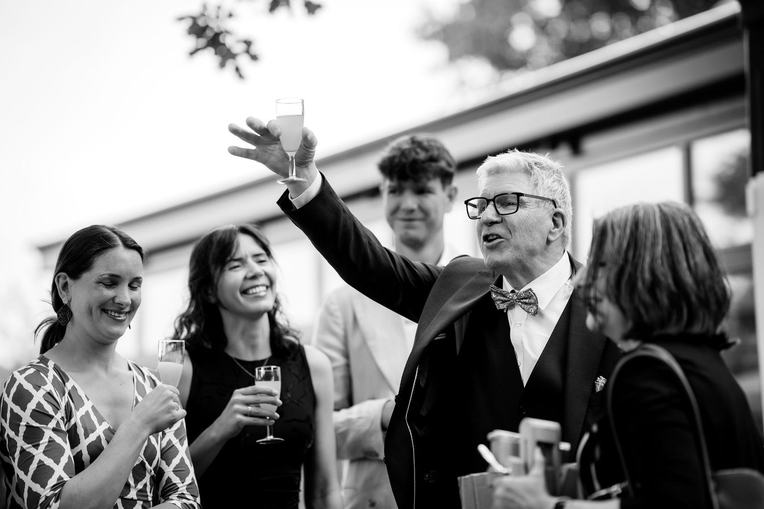Wedding guests raise glasses in a joyful toast outdoors in a black and white celebration photo.