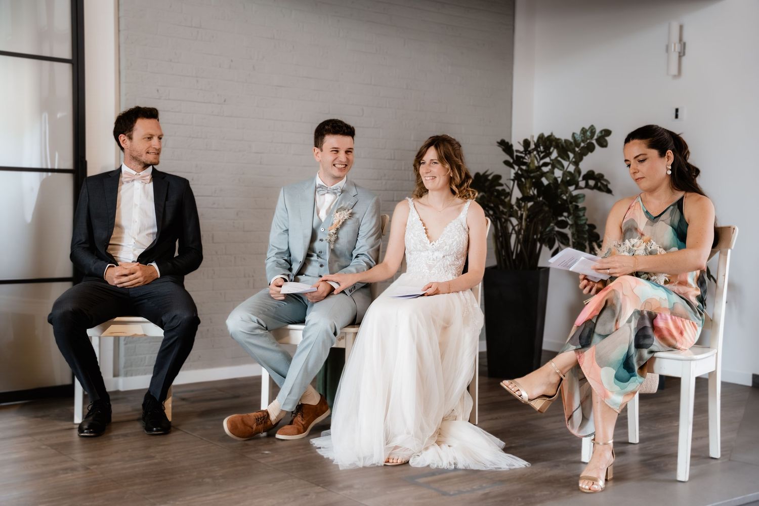 A bride and groom sit with two guests during a relaxed indoor wedding ceremony.