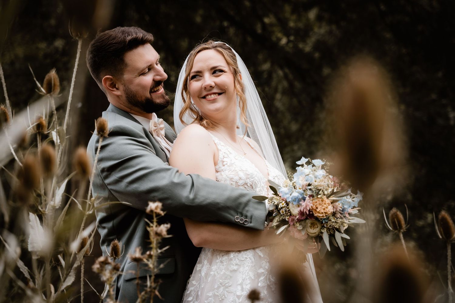 Bride and groom embrace outdoors among wildflowers, bride holding a pastel bouquet in a lace gown with veil.