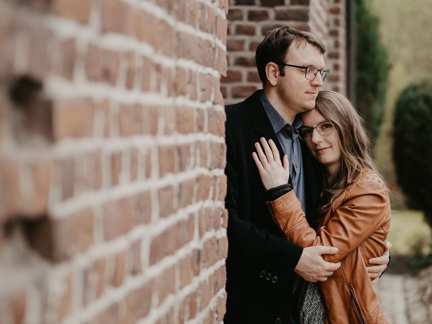 A couple embraces against a brick wall, with one wearing a brown leather jacket and the other in dark clothing.