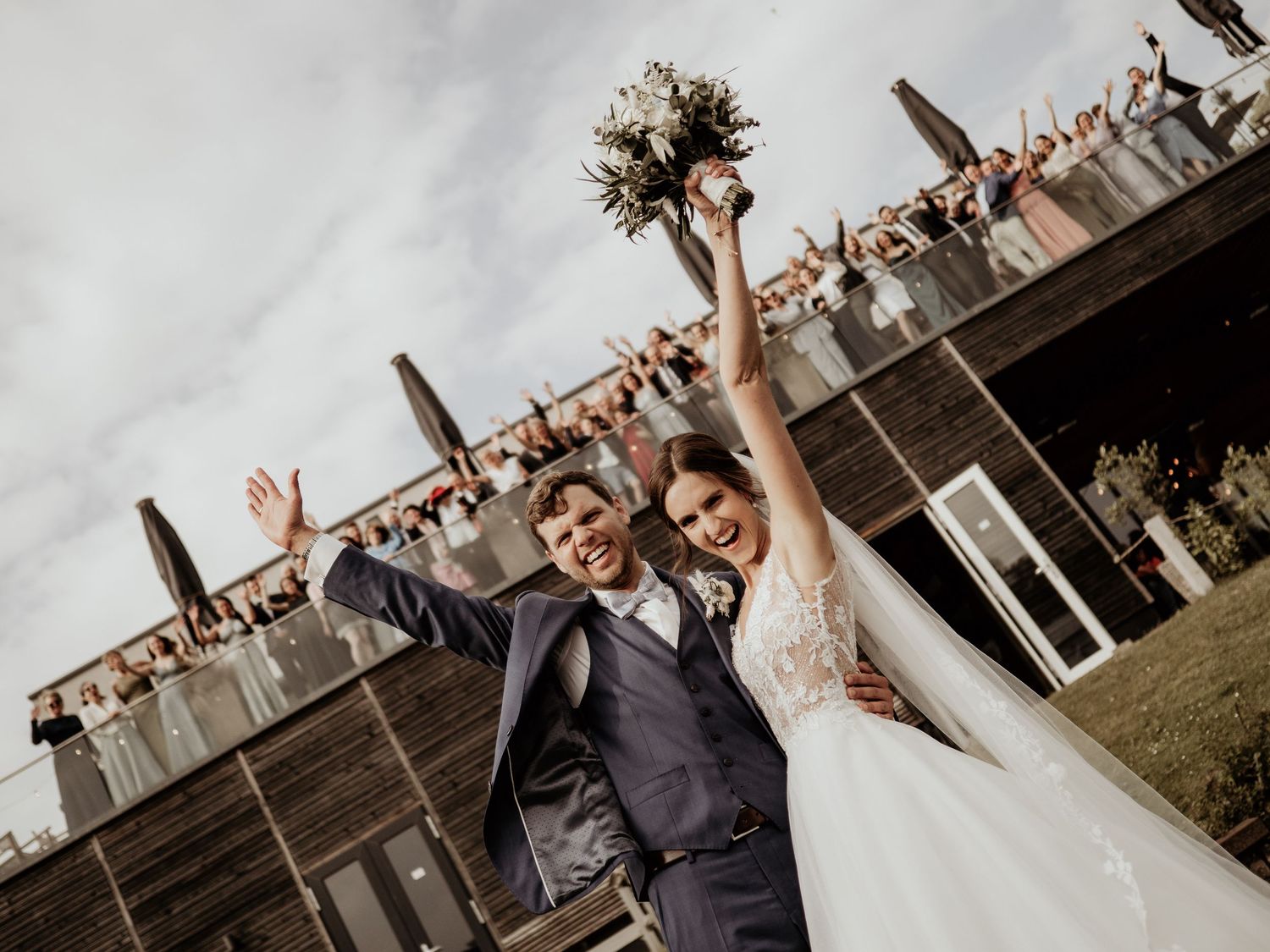 Wedding guests cheer and celebrate as the newlyweds share a joyful moment on an outdoor deck.