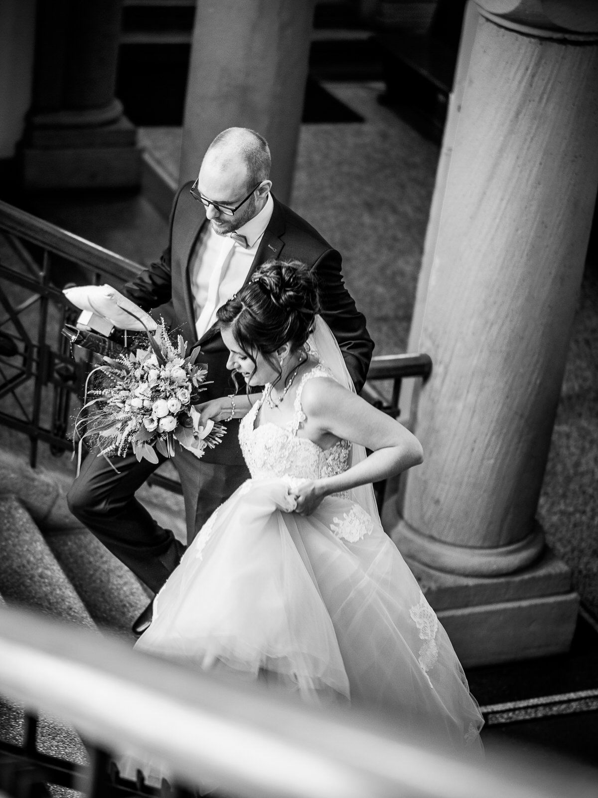 Black and white wedding photo of bride in white gown and groom descending stone steps with floral bouquet.