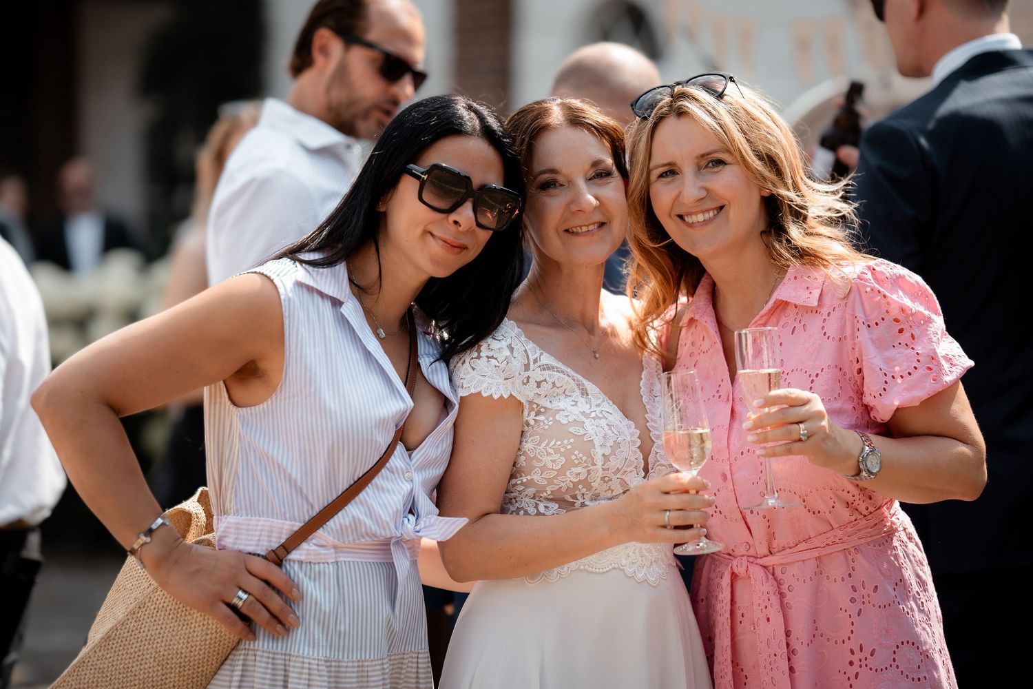 Bride posing with two smiling female guests outdoors, one holding champagne, at a sunny wedding reception.