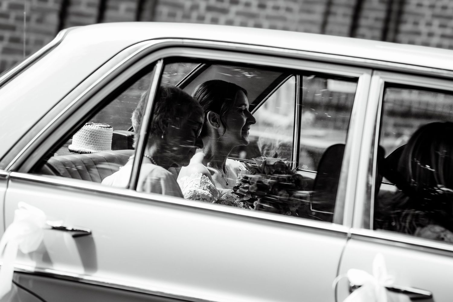 Black and white photo of a couple sharing a kiss inside a vintage car, captured through the window.