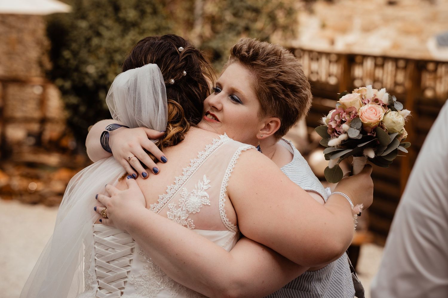 Emotional wedding moment as bride in lace gown receives warm hug from guest holding bouquet.
