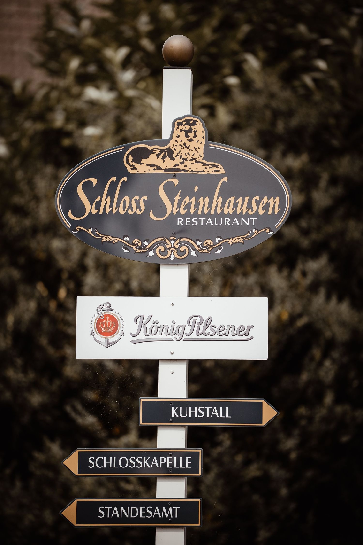 Sign post for Schloss Steinhausen Restaurant featuring König Pilsener and directional signs in Germany.
