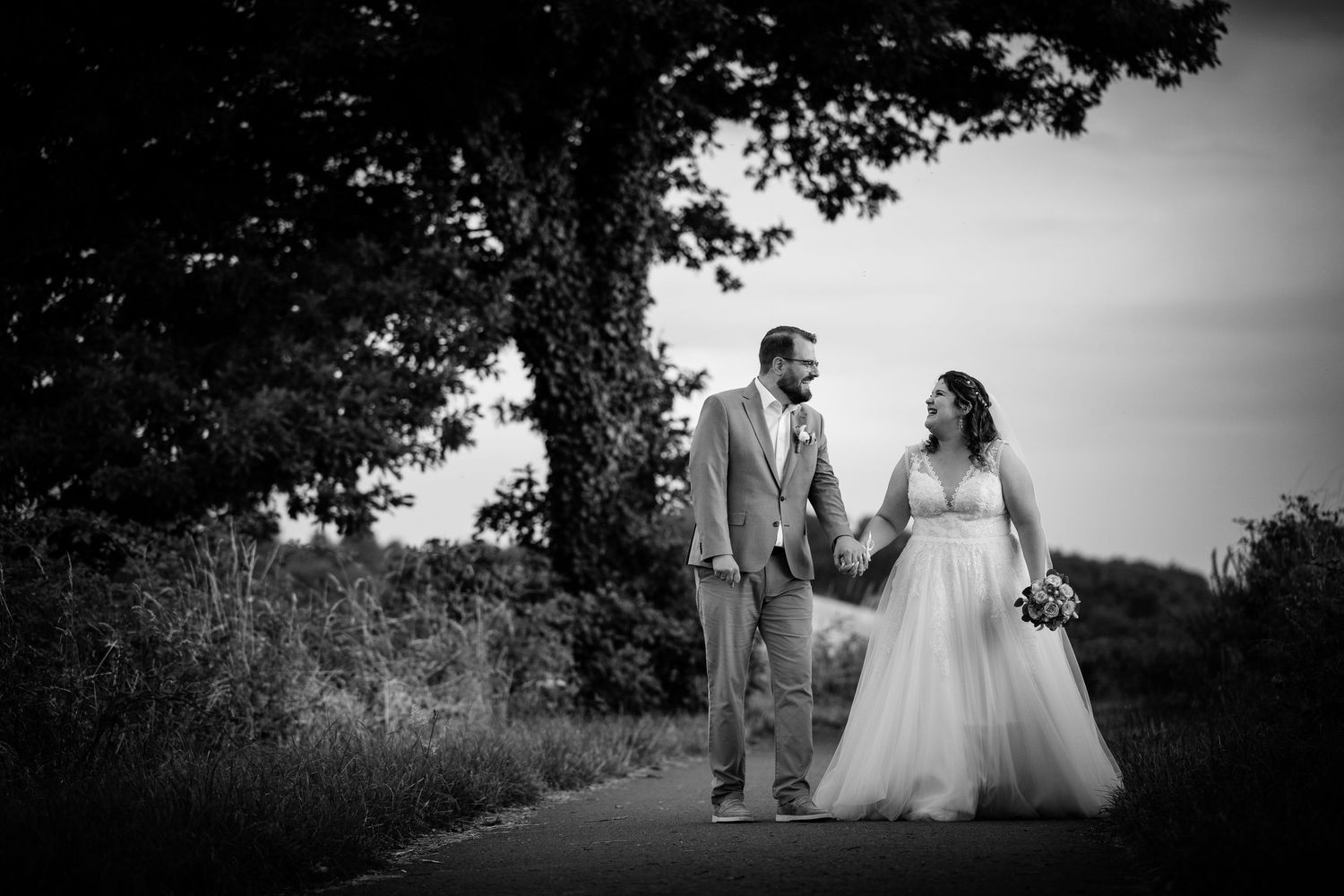 Black and white romantic photo of bride and groom walking hand in hand under a large tree.