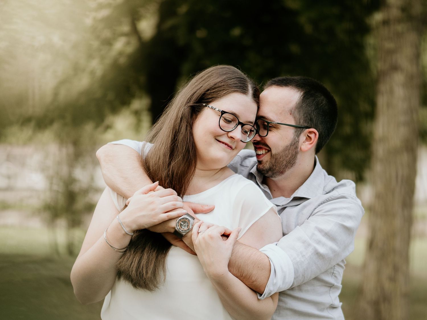 Couple embracing and smiling in a sunny park, woman wearing glasses with long hair.