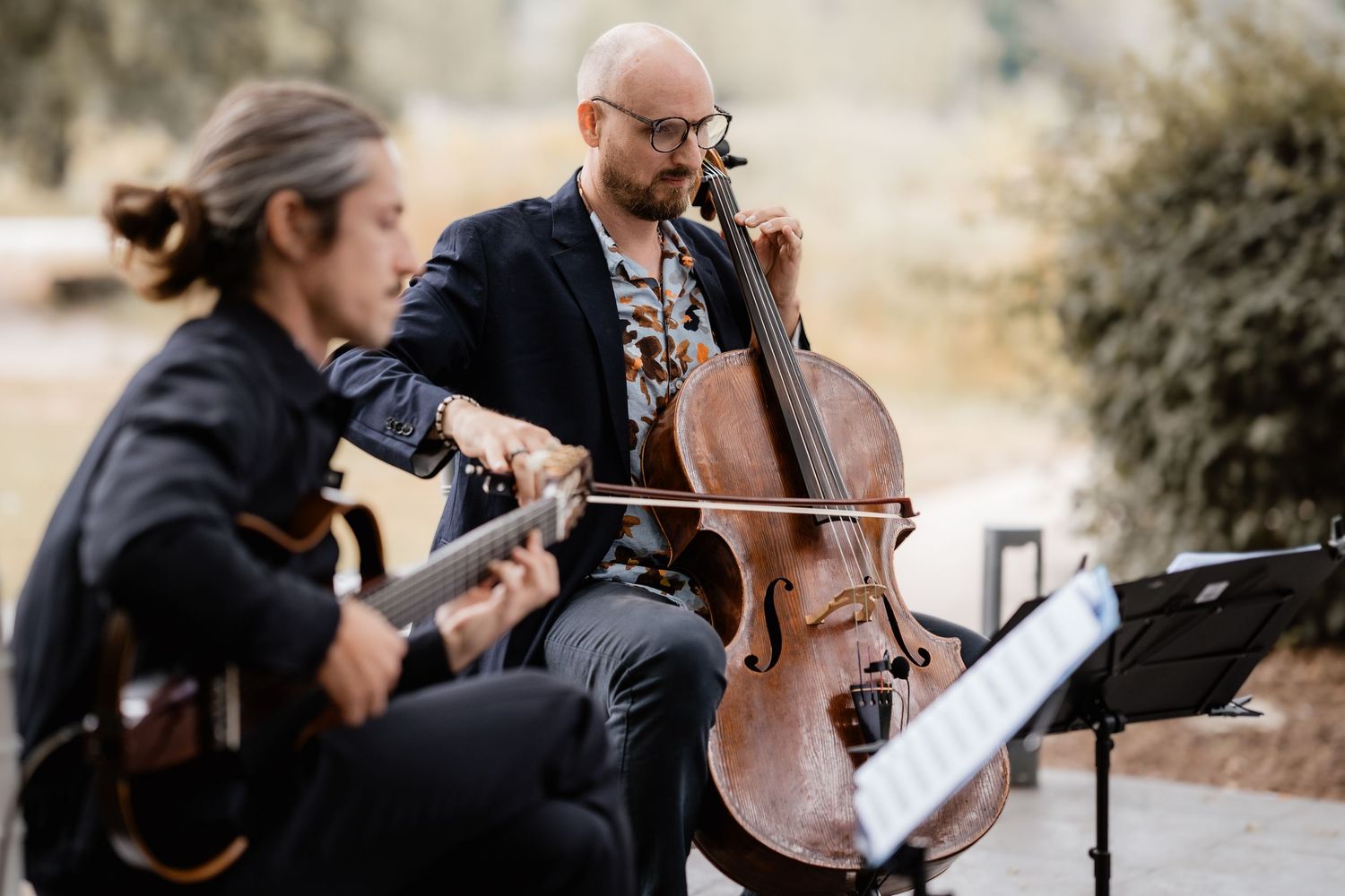 Two musicians playing cello and guitar outdoors at a wedding reception ceremony.