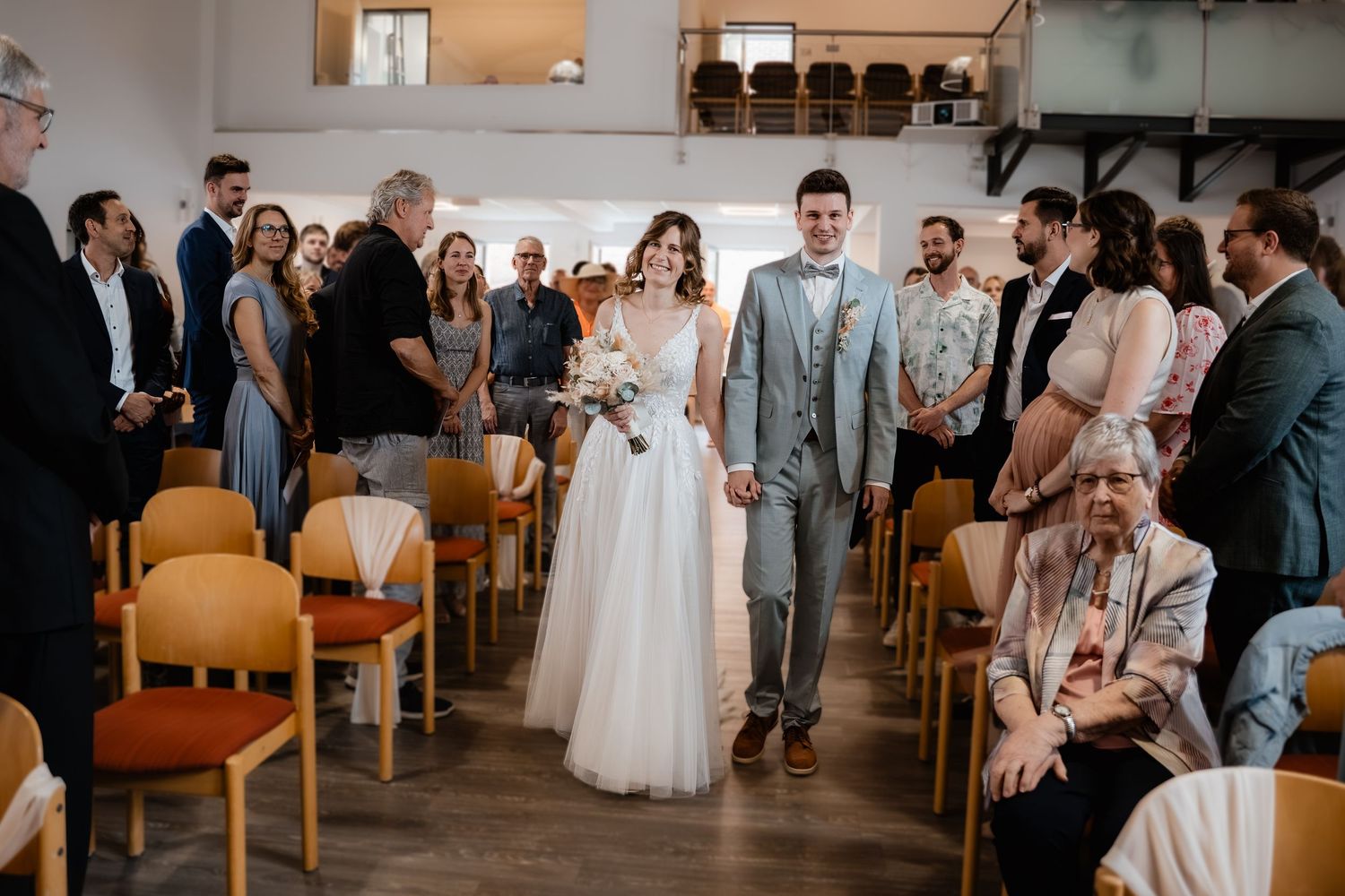 A bride and groom walk hand in hand down the aisle at their indoor wedding ceremony.