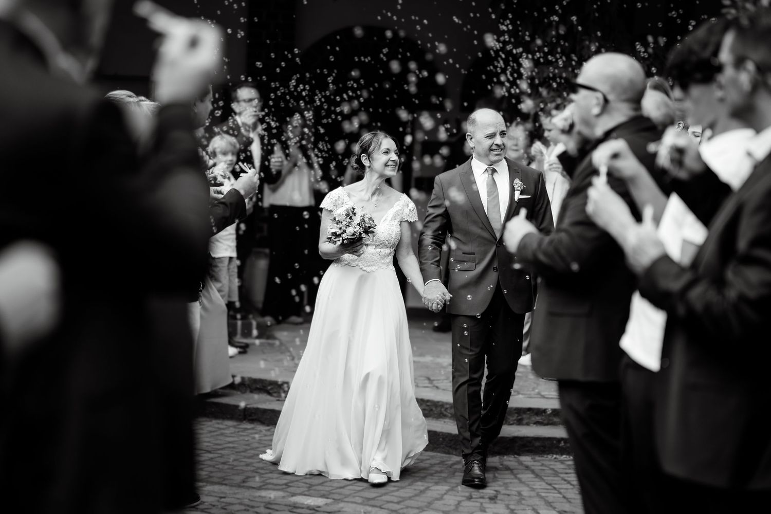 Black and white photo of newlyweds walking through confetti shower, smiling, surrounded by cheering guests.