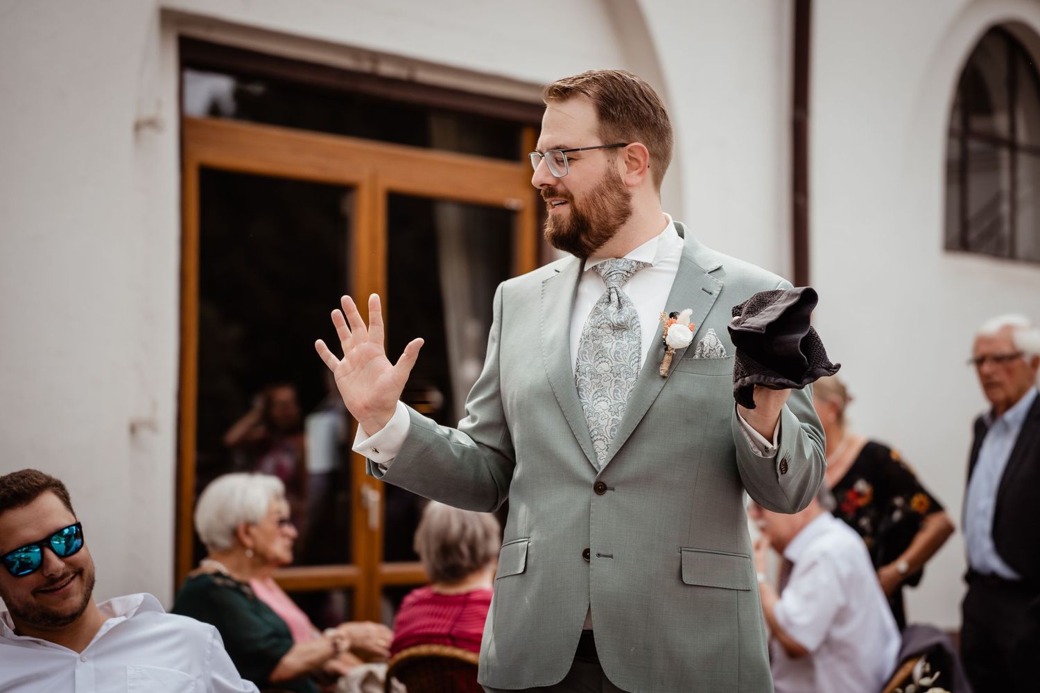 Groom in light gray suit waving at wedding guests outdoors, holding jacket, with crowd seated in background.