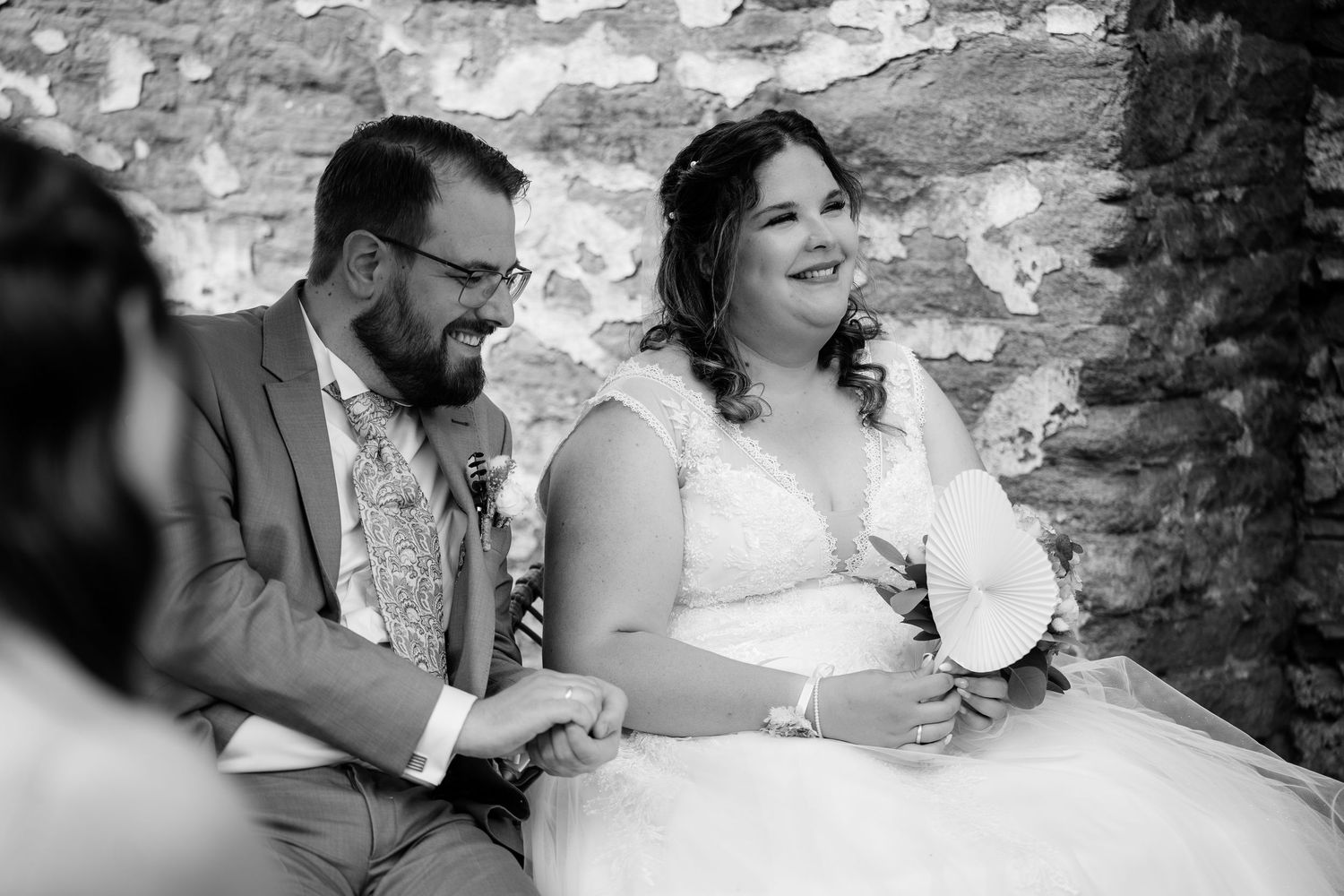 Bride and groom laughing joyfully during wedding ceremony seated against stone wall in black and white photo.