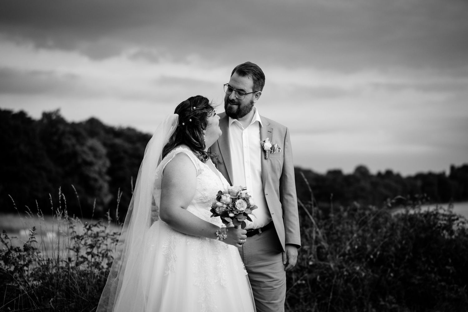 A bride and groom share a tender moment outdoors in a black and white wedding portrait under dramatic skies.