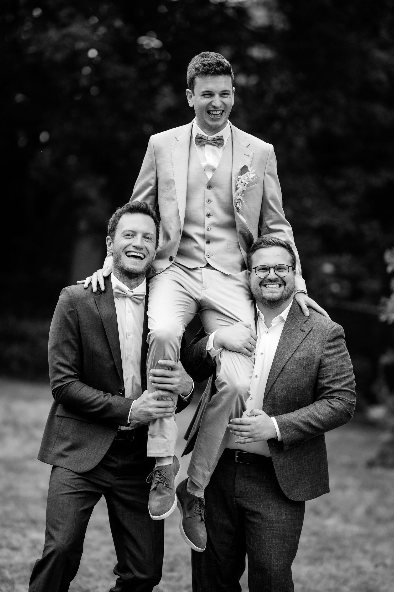 Groom lifted by two laughing groomsmen in black and white wedding photo.