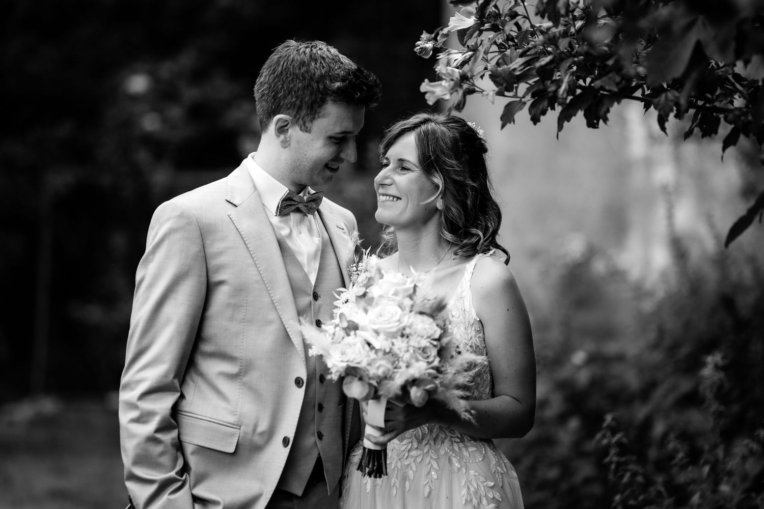Black and white photo of bride and groom smiling at each other under leafy branches.