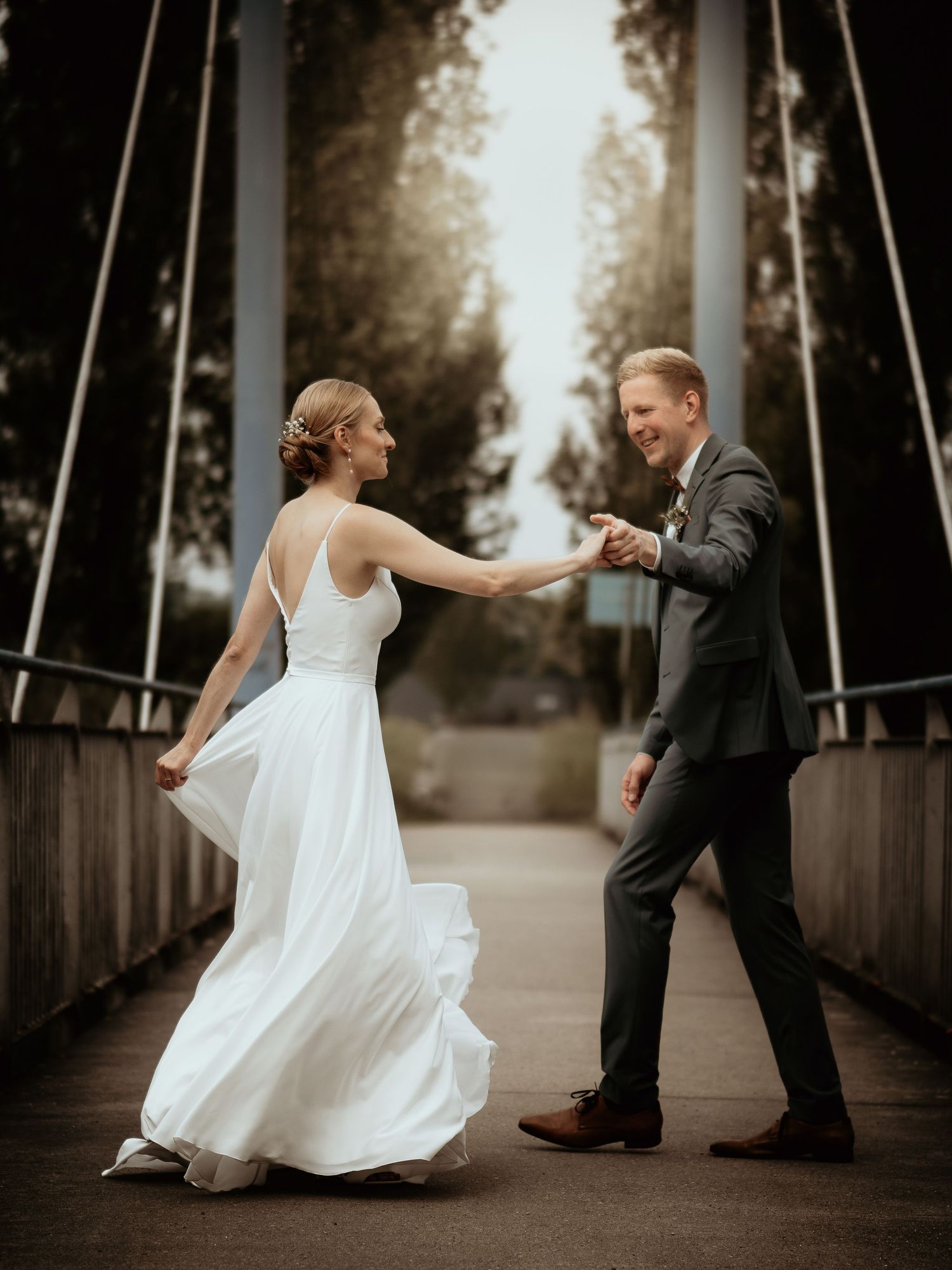 Bride and groom dancing joyfully on a wooden bridge, bride in flowing white gown, groom in dark suit.