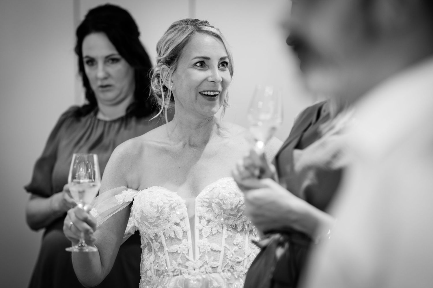Bride in lace gown reacting with surprise while holding champagne glass at reception.