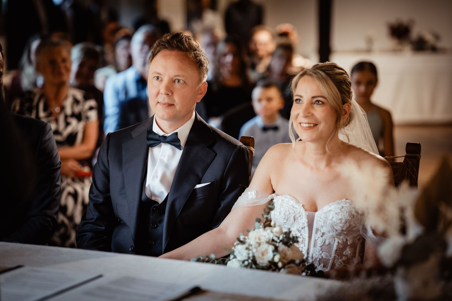 Bride and groom smiling during wedding ceremony seated at table with bouquet.