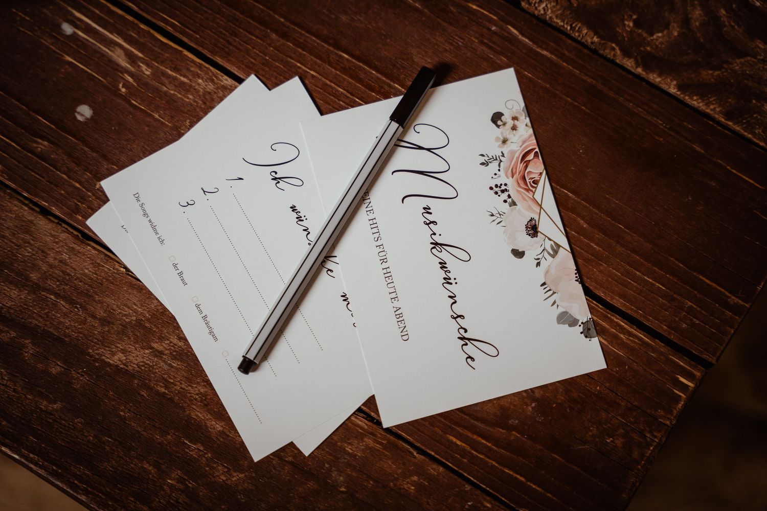 Elegant wedding stationery cards with calligraphy script and floral design, paired with a black pen on rustic wood.