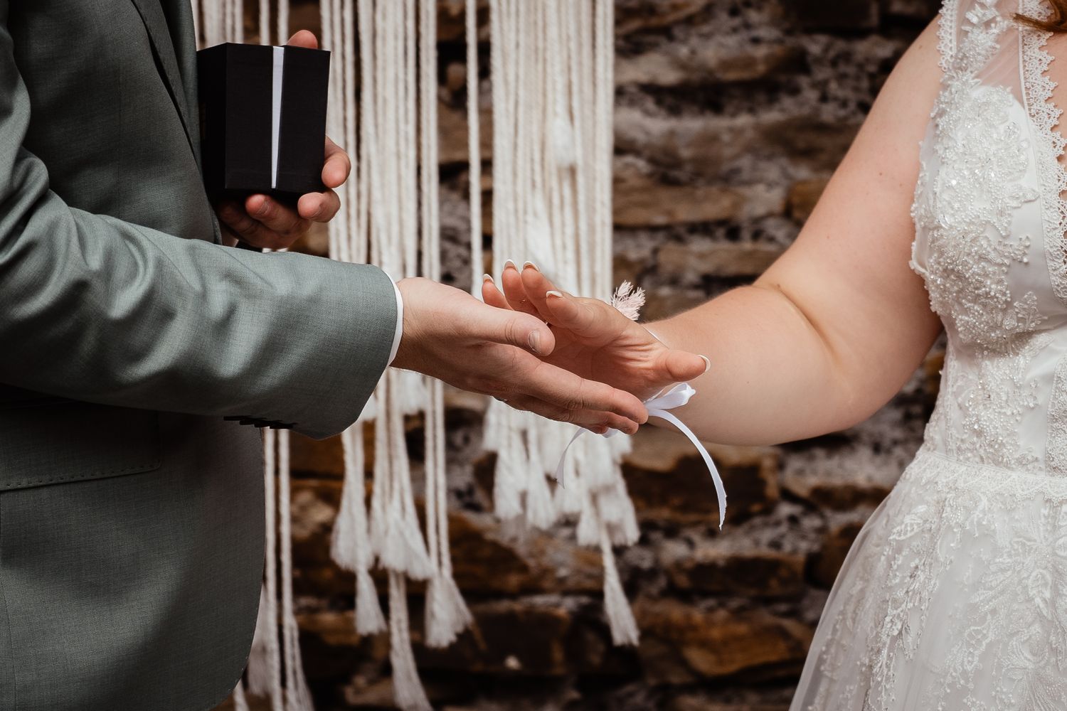 Groom placing wedding ring on brides hand during ceremony with macrame backdrop.