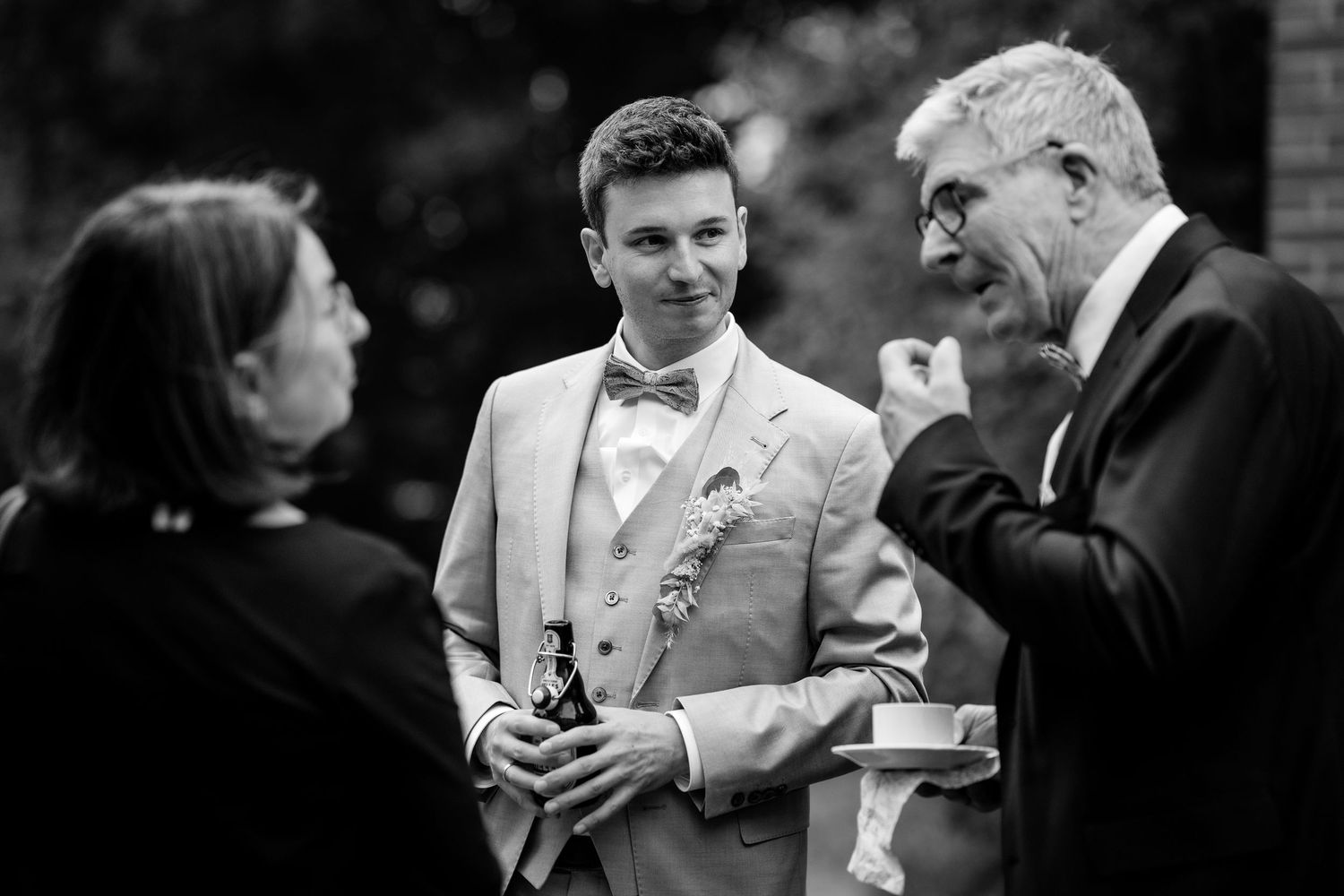 Groom in a light suit with bow tie smiling while chatting with guests at a wedding reception.