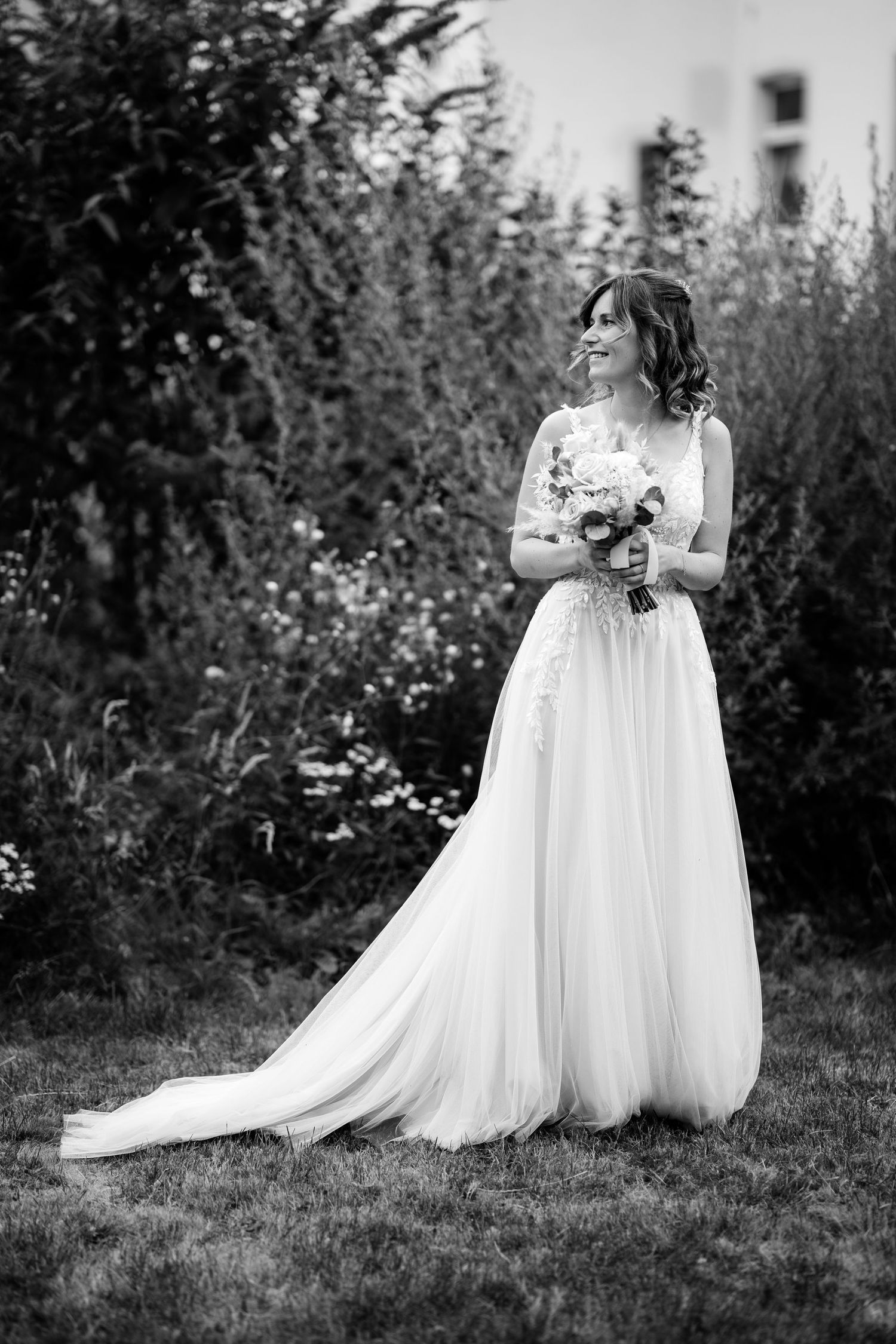 Elegant bride in a flowing white lace gown holding a bouquet, standing in a lush garden in black and white.