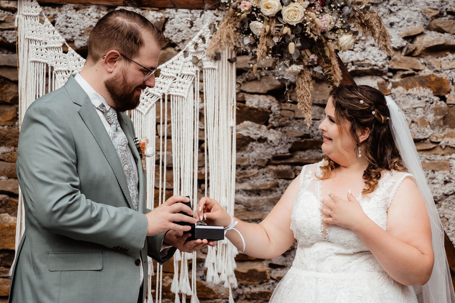 Groom in sage suit places ring on laughing bride's finger at boho stone wall ceremony backdrop.